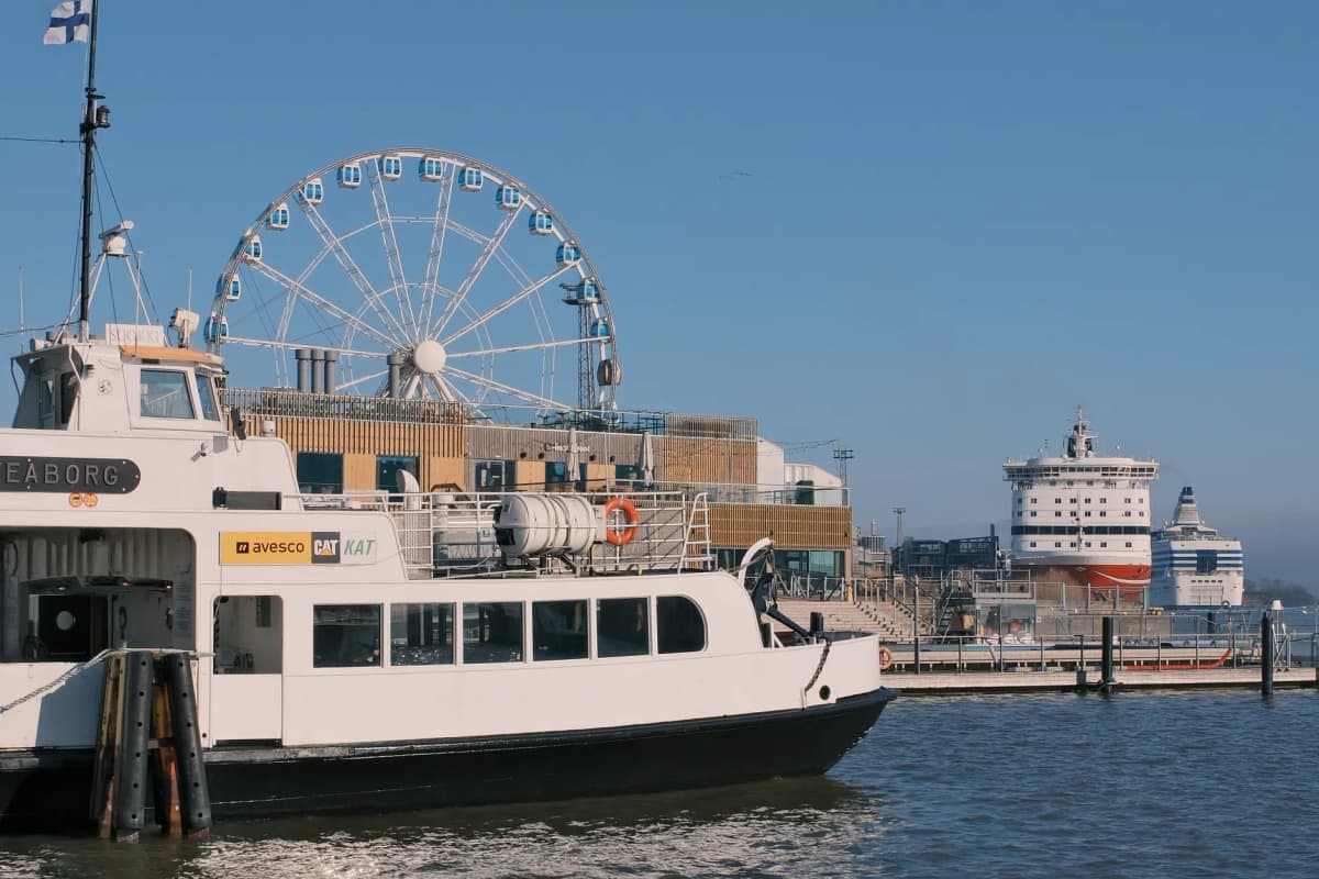 City harbor scene featuring ferris wheel, ferry, and cruise ship under clear blue sky.